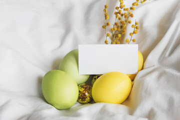 painted easter yellow and green chicken and quail eggs, spring mimosa sprig, greeting white card blank for your text on white cloth, easter holiday celebration concept 