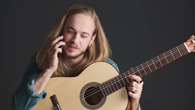 Young long haired blond man with guitar talking with friend on smartphone waiting for rehearsal to start isolated on black background