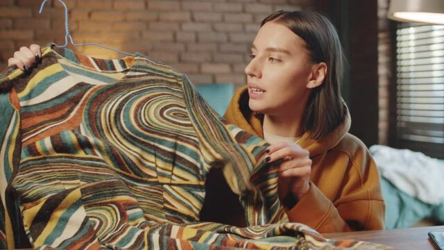 Young beautiful woman showing dress and talking at camera while video calling or recording fashion vlog at home