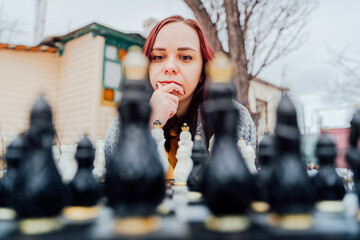 Young woman playing chess in yard. Female wrapped in grey plaid sitting on street playing in board game in winter season.
