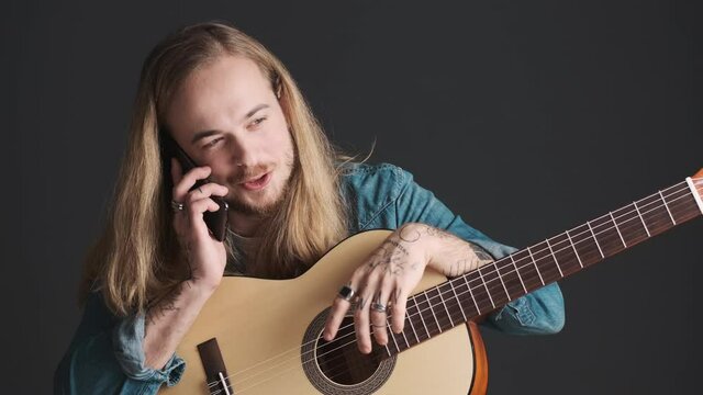 Long haired blond guy with acoustic guitar talking with friend on smartphone after rehearsal in studio isolated on black background