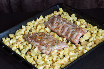 Strips of fresh pork rib bathed in a sauce of garlic, oil, vinegar, and oregano surrounded by potatoes on a baking sheet. Fresh food and raw meats on a wooden table.