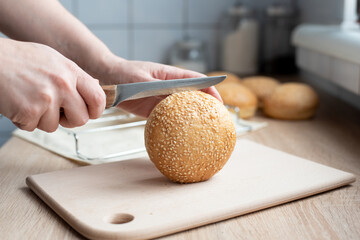 Woman's hand cutting a sesame seed bun for a hamburger with a knife in the kitchen, cooking at home.
