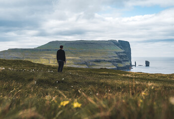 Fototapeta premium Young man tourist standing in front of Risin and Kellingin rocks in the sea as seen from Tijornuvik bay on Streymoy on the Faroe Islands, Denmark, Europe