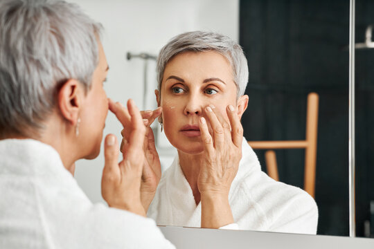 Mature Woman Applying Moisturizer In Bathroom. Senior Female In Bathrobe Doing Skincare Routine.