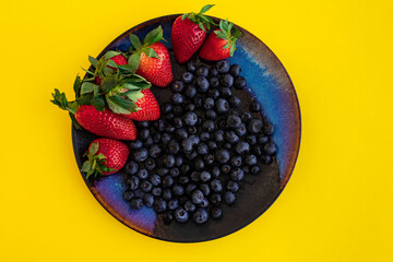 Fresh ripe large strawberries and blueberries on a colored plate on a yellow background, top view.