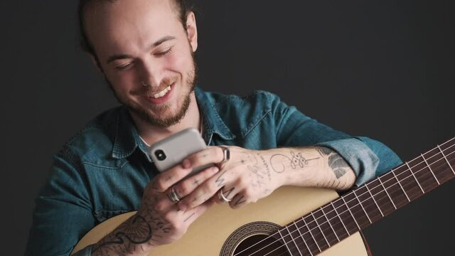 Young bearded man with guitar chatting with friend on smartphone waiting for rehearsal to start over black background. Modern technology concept