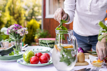 Woman putting mint herb into cold lemonade in pitcher. Preparing non-alcoholic drink for garden...
