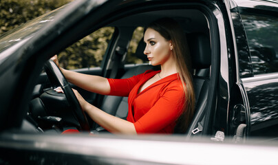 Obraz na płótnie Canvas A beautiful young girl in a red overalls sits behind the wheel of a black car on an empty road in the forest