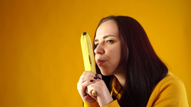 Portrait of young woman with banana on yellow background. Close up of female in yellow hoodie plays with fruit, imagining it as weapon.