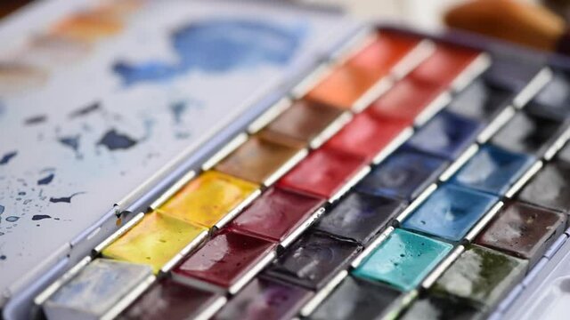 A set of bright saturated watercolors and brushes for painting in the artist's workshop. Mixed shades of paints on a plastic palette. Drawing and creative tools close-up. The camera is in motion.