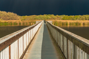 Obraz premium Lake and long wooden boardwalk through the lake with dark blue sky on background, few minutes before the storm. Oso Flaco Lake Natural Area in Oceano, California