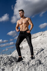 Handsome bodybuilder standing on white mountain and looking away. Man with muscular body type in wirstwatch holding sunglasses. Attracive man with naked abs.