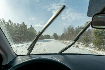 The view from the car with working windshield wipers on a snowy road in the forest. Snowstorm in sunny weather