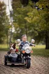 cute boy enjoying in autumn park, driving electrical motorcycle toy with sidecar and his dog in it