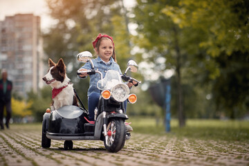 female preschooler driving electrical motorcycle toy with sidecar and her dog in it, smiling