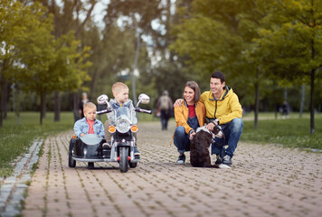 young caucasian parents  playing in park with their sons in motorcycle toy and a dog