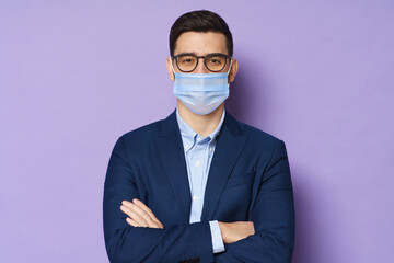 Young business man wearing glasses and medical mask, standing with crossed arms isolated on purple...