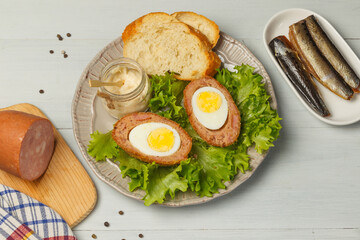 Scottish-style eggs cut in half on a plate with lettuce leaves, baguette slices and sauce. On a light wooden table, close up with space