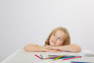 A small cute schoolgirl girl with blonde hair draws an inscription in an album with colored pencils on a light background