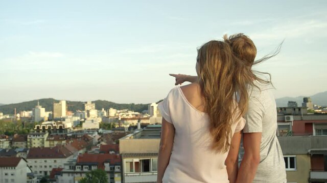 SLOW MOTION, CLOSE UP, DOF: Couple standing on the edge of rooftop with great view, looking at beautiful colorful city. Boyfriend showing his home to girlfriend pointing with finger into the distance