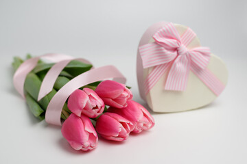 Fototapeta na wymiar Beautiful tulips of pink color on a white background near a gift box in the shape of a heart with a ribbon and a beautifully tied pink bow. The concept of a gift and surprise for a loved one.
