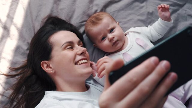 Top view of cute young mother and beautiful newborn baby making selfie or video call to relatives lying on comfortable grey bed at home. Technology, new generation, connection, parenthood concept