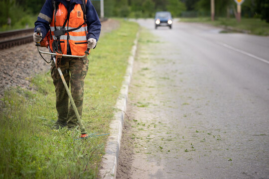 Front View Of A Worker In Protective Clothing With A Petrol Lawn Mower On Wheels Walking On A Lawn With Grass Along The Roadway. A Man Mows Grass With Dandelions By The Road In The City.