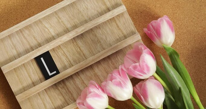 Women's hands fold the word love from letters in a wooden box. Nearby is a bouquet of pink tulips. Top view. Flat lay.