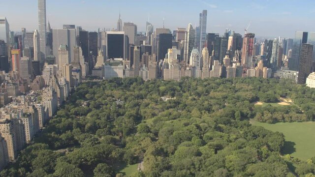 AERIAL ESTABLISHING SHOT: Flying above Central Park along 5th Avenue and towards Downtown Manhattan in sunny New York City. Tall glassy skyscrapers and condominium buildings overlooking Central Park