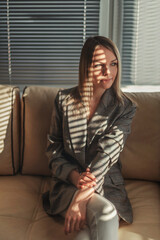 A beautiful blonde girl in a plaid jacket sits on a light sofa in the sun