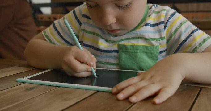 A portrait of a child sits at a table at home and draws on his tablet with a special pen, a preschooler uses a special tablet for drawing.