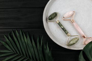 Facial roller from crystal rose quartz  and massage tool jade Gua sha on the tray  on black background with palm leaves. Tools for anti-age massage, natural face lifting  at home. Dark mood style