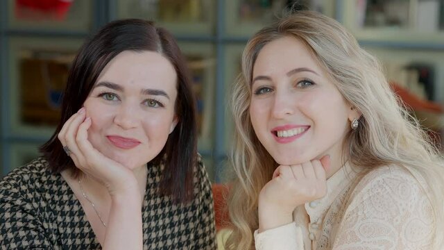 Two positive young women with toothy smile looking at each other and posing. Portrait of happy carefree relaxed Caucasian female friends in cafe or restaurant indoors. Lifestyle and beauty concept.