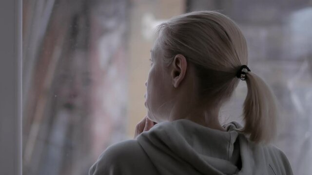 A woman brushes her teeth while holding a toothbrush in her left hand, standing by the window