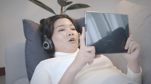 Happy cheerful Asian woman wearing wireless headphones watching to the movie from a tablet in relax time at home lifestyle and pleasure concept