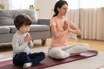 Mommy practicing, teaching child to meditate. Fit mom with little son doing yoga exercise sit on yoga mat on floor at home. Calm mother and kid sitting in lotus pose. Stress relieve, wellness concept.