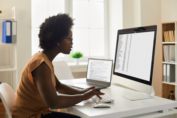 Employee using office computer. African-American woman sitting at desk, looking at desktop PC screen, doing research, studying data reports, working with business documents and online spreadsheets - Powered by Adobe