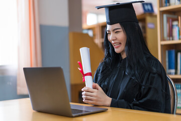 A young Asian university graduate woman in graduation gown expressing joy and excitement to celebrate her education achievement in front of a laptop making a remote video call to her parents at home