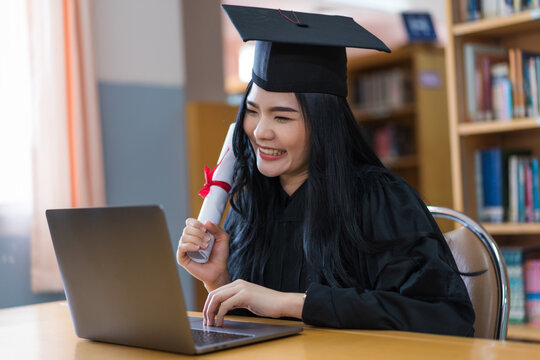 A young Asian university graduate woman in graduation gown expressing joy and excitement to celebrate her education achievement in front of a laptop making a remote video call to her parents at home