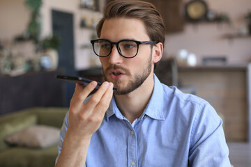 Confident business man in glasses holding smartphone near mouth for recording voice message or...
