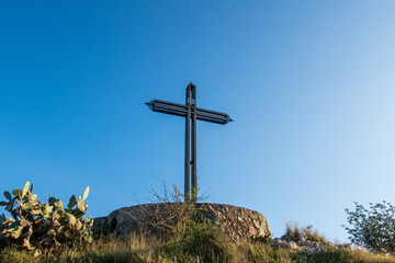 Iron cross on the mountain of El Rabat, in Valencia (Spain), La Safor region, on a day with blue skies, and surrounded by vegetation and flowers.