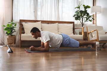 Side view of Black African American man looking at digital tablet and doing plank exercise at home....
