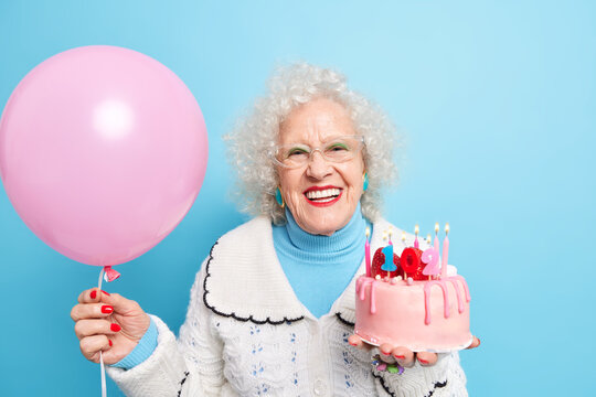 People old age partying and holidays concept. Positive beautiful grandmother dressed in neat clothes celebrates her 102nd birthday holds inflated balloon and tasty cake isolated over blue background