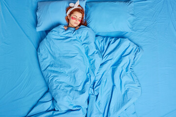 Overhead shot of redhead young European woman sleeps peacefully in bed sees sweet dreams applies collagen patches under eyes poses under soft blanket poses at bedroom. People rest sleeping concept