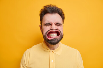 Headshot of bearded super crazy adult man clenches teeth closes eyes has wide opened mouth wears casual shirt isolated over vivid yellow background. People emotions face expressions concept.