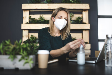 pretty, young blond businesswoman is sitting in a sustainable, ecological office wearing a protective mask and disinfecting her hands because of corona