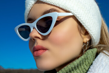 Portrait of a young girl in winter clothes on a background of a blue sky in winter clothes and fashionable sunglasses