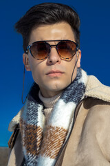 Portrait of a young guy in winter clothes against a blue sky in winter clothes and fashionable sunglasses