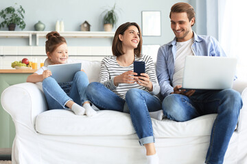 Father, mother and daughter using electronic devices sitting on sofa at living room.
